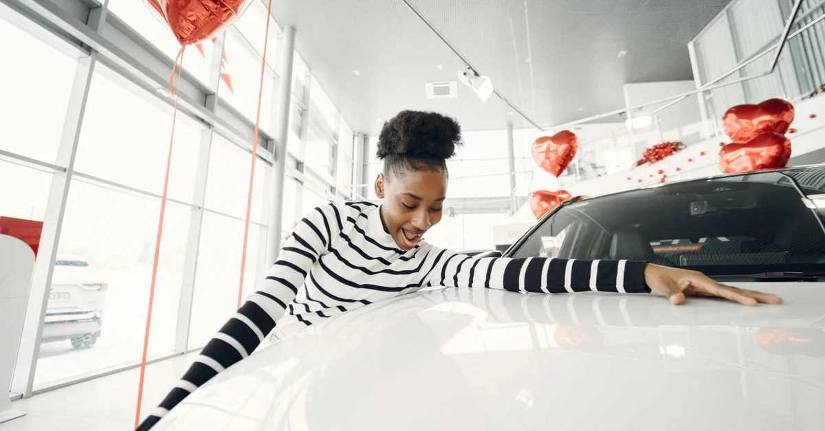 Femme excitée étreignant sa nouvelle voiture chez un concessionnaire, entourée de ballons rouges.