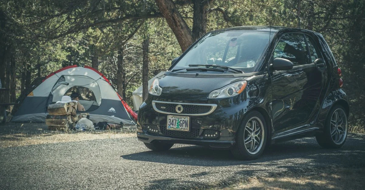 Voiture Smart garée à côté d'une tente dans un camping boisé, idéale pour les aventures en plein air.
