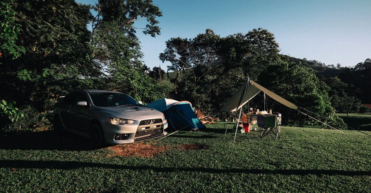 Scène de camping paisible avec une voiture garée et une tente au milieu d'une végétation luxuriante sous un ciel dégagé.